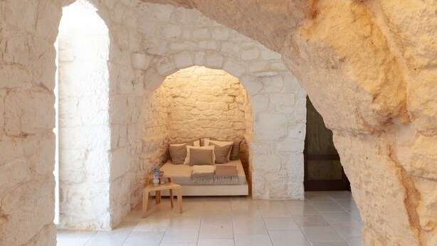 Quarto com paredes de pedra, uma cama, uma mesinha e um banco.