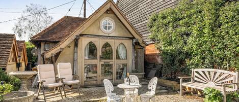 A rustic building with a triangular roof, a stone fountain, and outdoor seating with chairs and a table.
