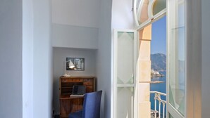 Quarto com escrivaninha de madeira, um laptop, uma cadeira, luminária de mesa e um quadro na parede. Há uma varanda com vista para o mar e uma montanha.