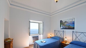 Une chambre avec deux lits, une fenêtre donnant sur la mer, un tableau au mur et un plafond décoratif.