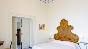Een slaapkamer met een groot, sierlijk bed, een deur naar een andere kamer en een klein tafeltje met een lamp.