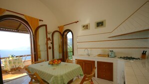 A dining area with a table set for two, a kitchen with a gas stove, and a view of the sea through an open archway.