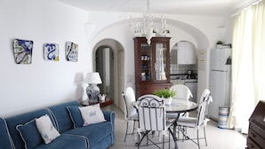 Ein modernes Wohnzimmer mit einem blauen Sofa, einem Esstisch mit Stühlen und einer Küchenzeile mit weißen Schränken und einem Kronleuchter.