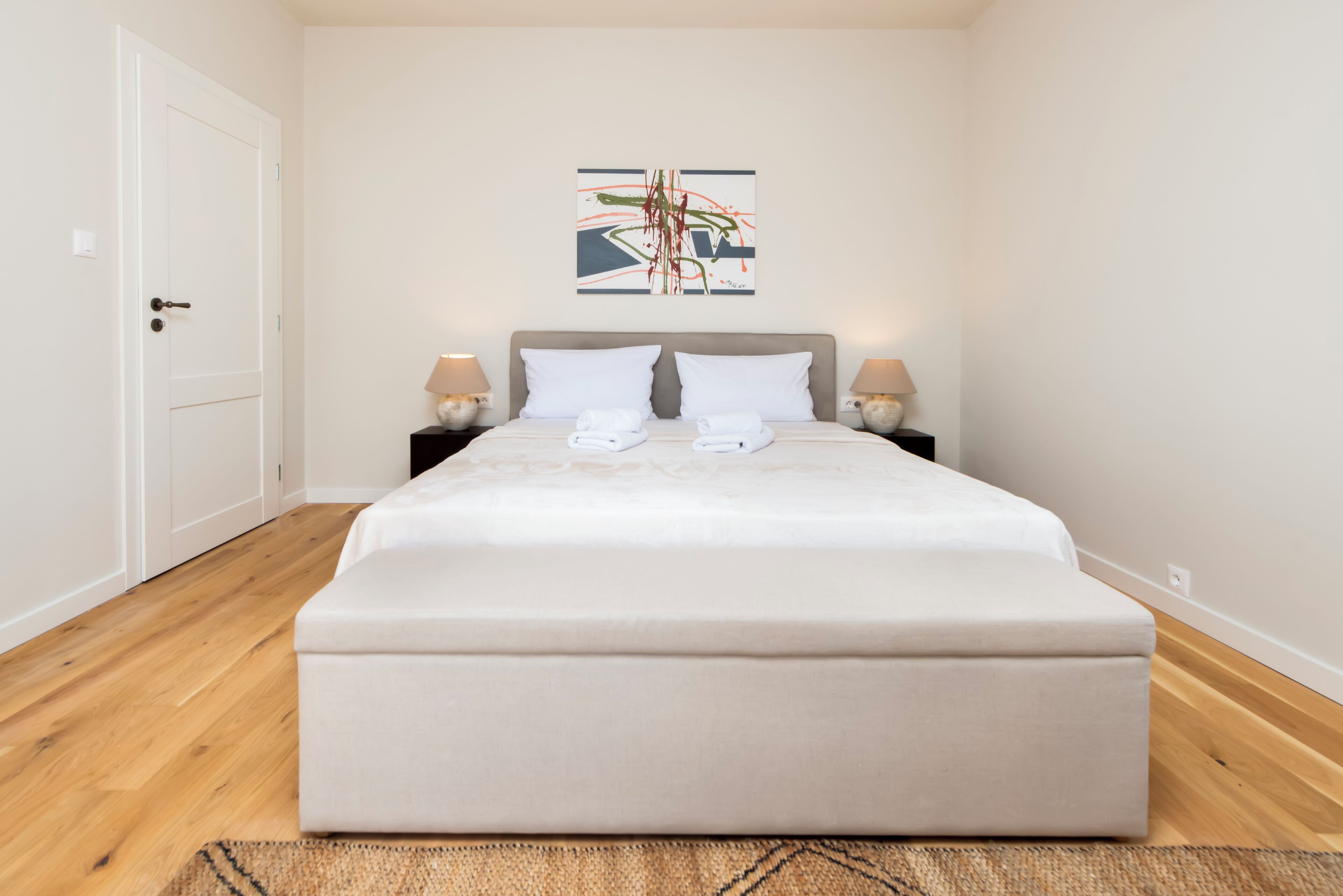 A neatly made bed with white linens and pillows, a bedside table with a lamp, and a wall-mounted abstract art piece.