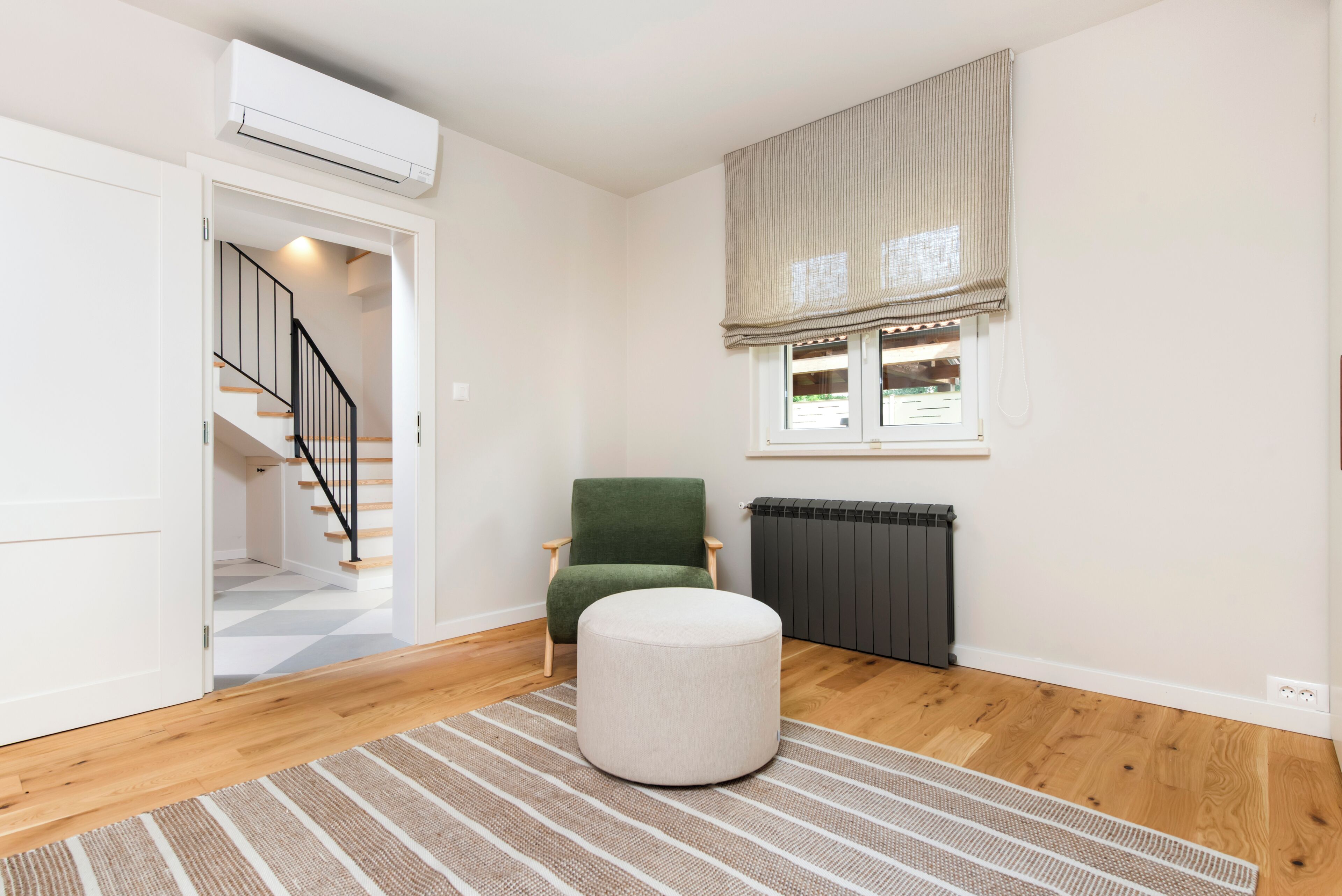 Una habitación con suelo de madera, una silla verde, un reposapiés blanco, un radiador y una ventana con persiana.
