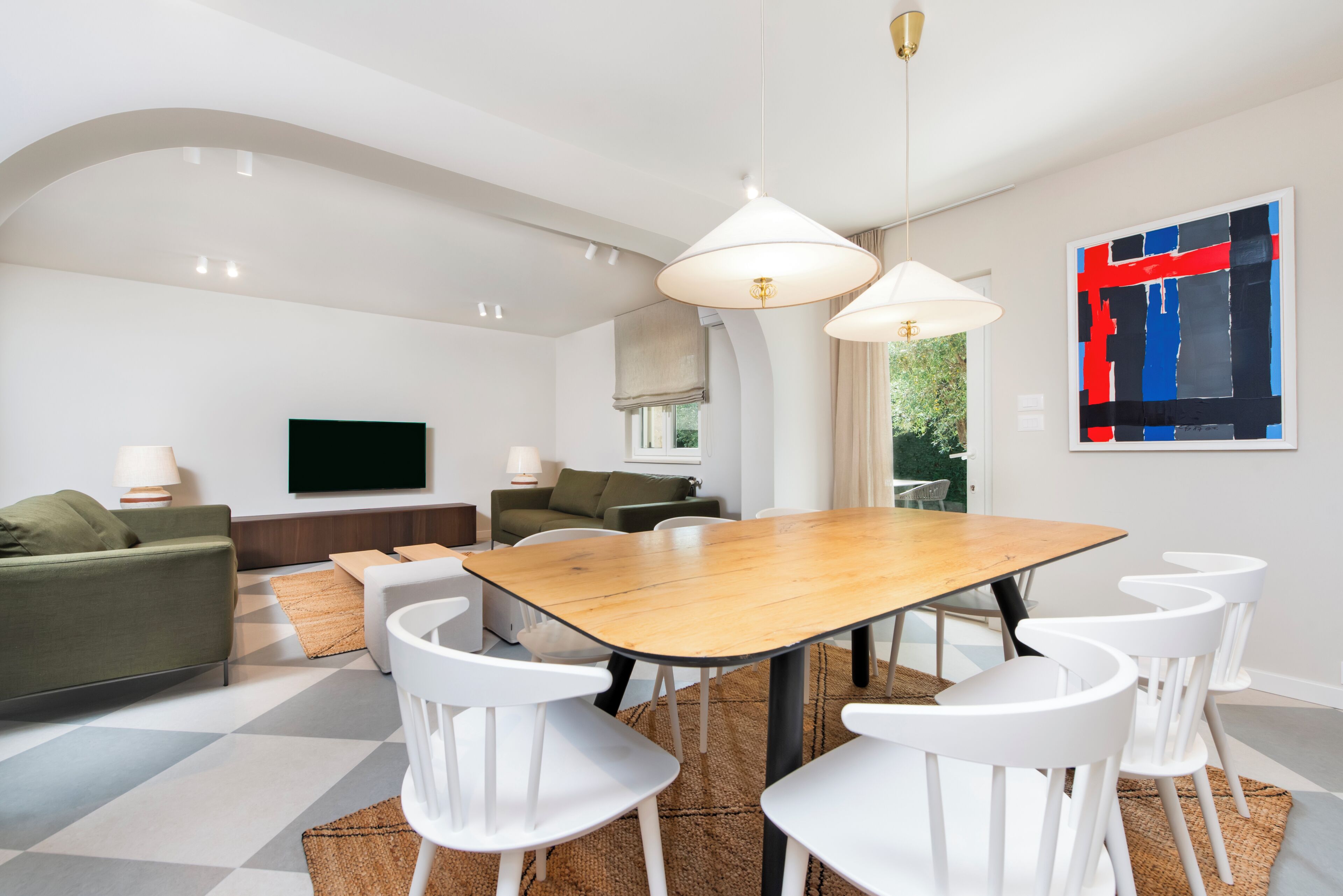 Una sala de estar moderna con comedor, que presenta una gran mesa de madera, sillas blancas y un televisor en la pared.
