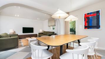Una sala de estar moderna con comedor, que presenta una gran mesa de madera, sillas blancas y un televisor en la pared.