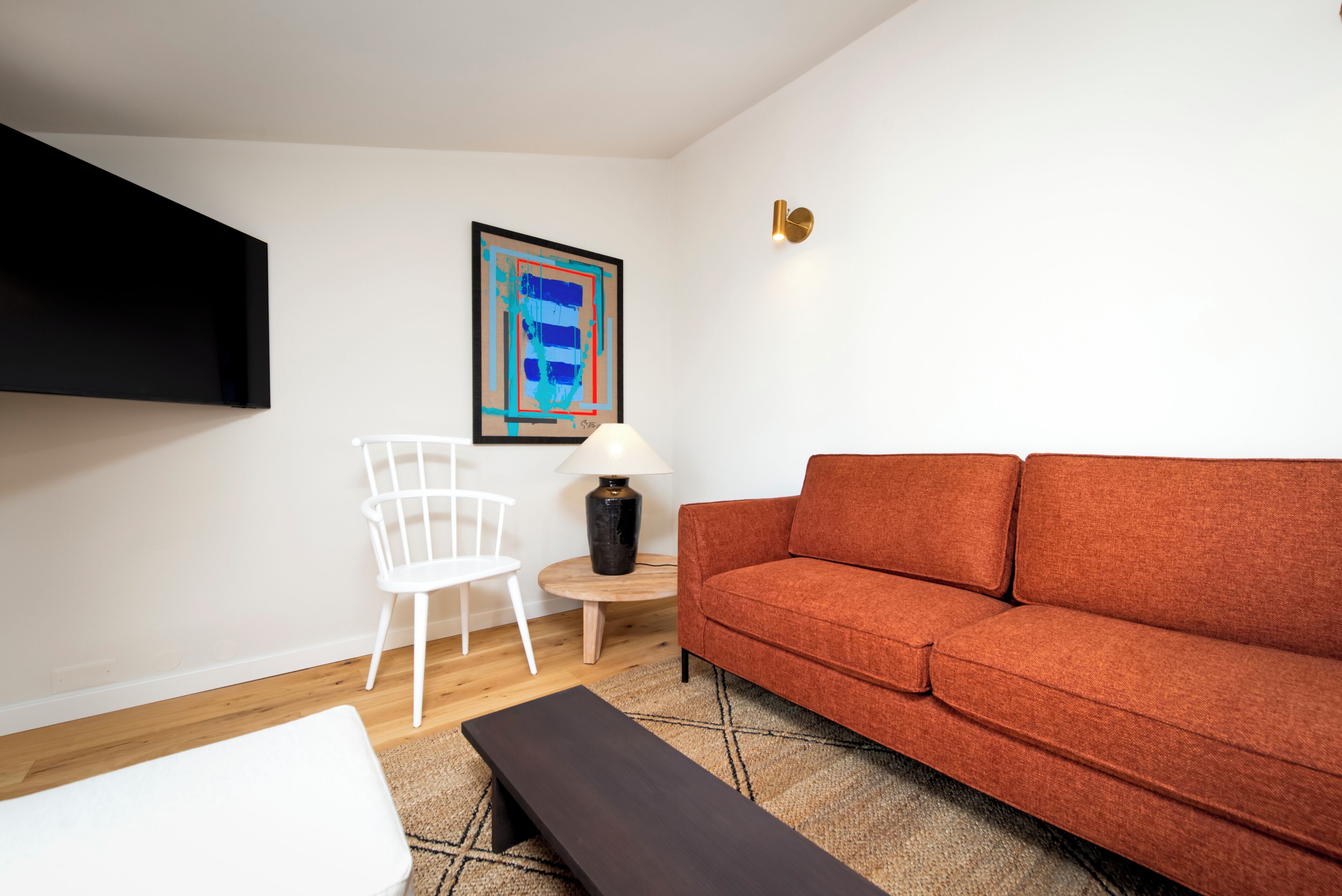 Uma sala de estar com um sofá, uma cadeira branca, uma mesinha com abajur e um quadro na parede.