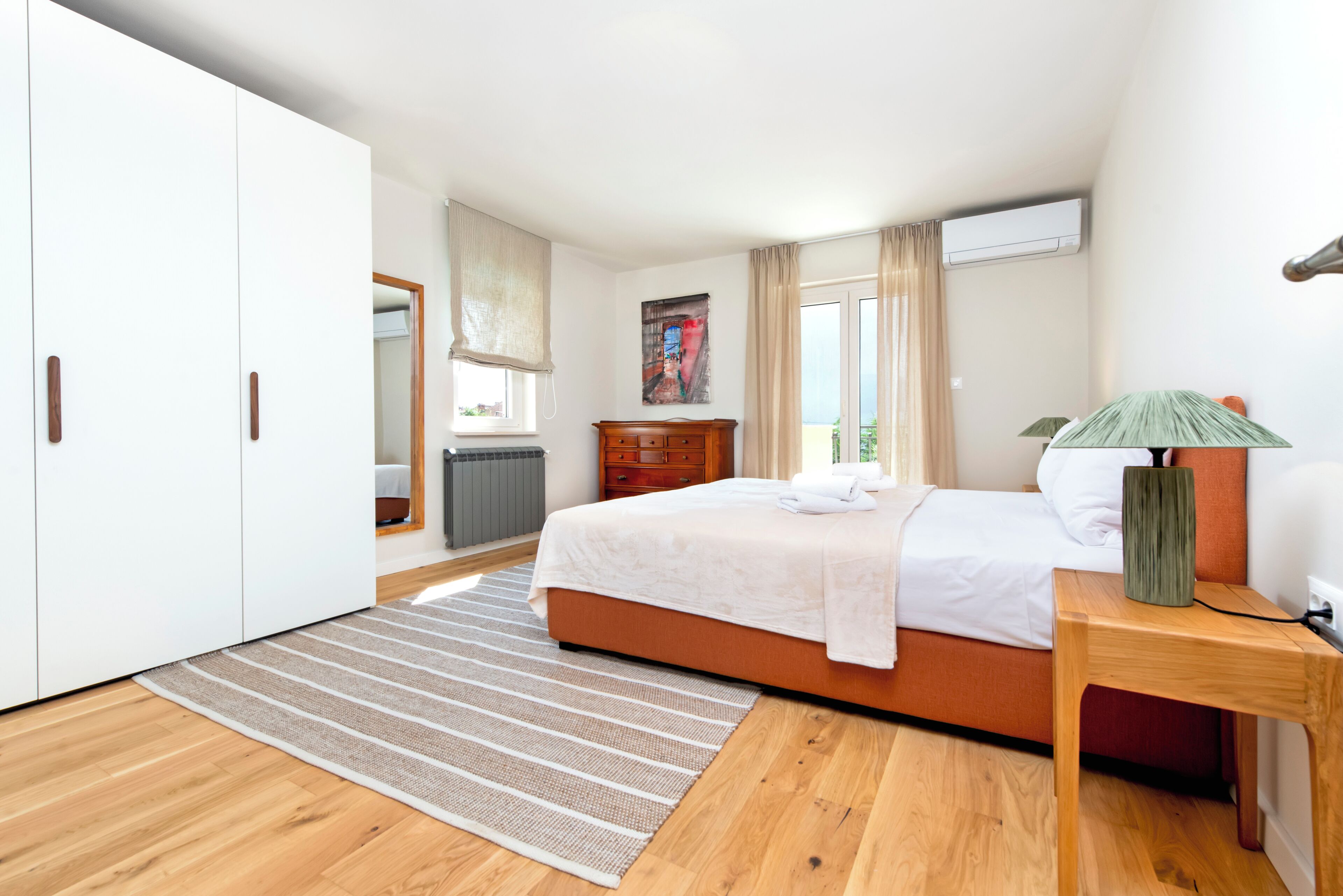 A bedroom with a bed, a wooden nightstand, a dresser, a window with curtains, and an air conditioning unit.