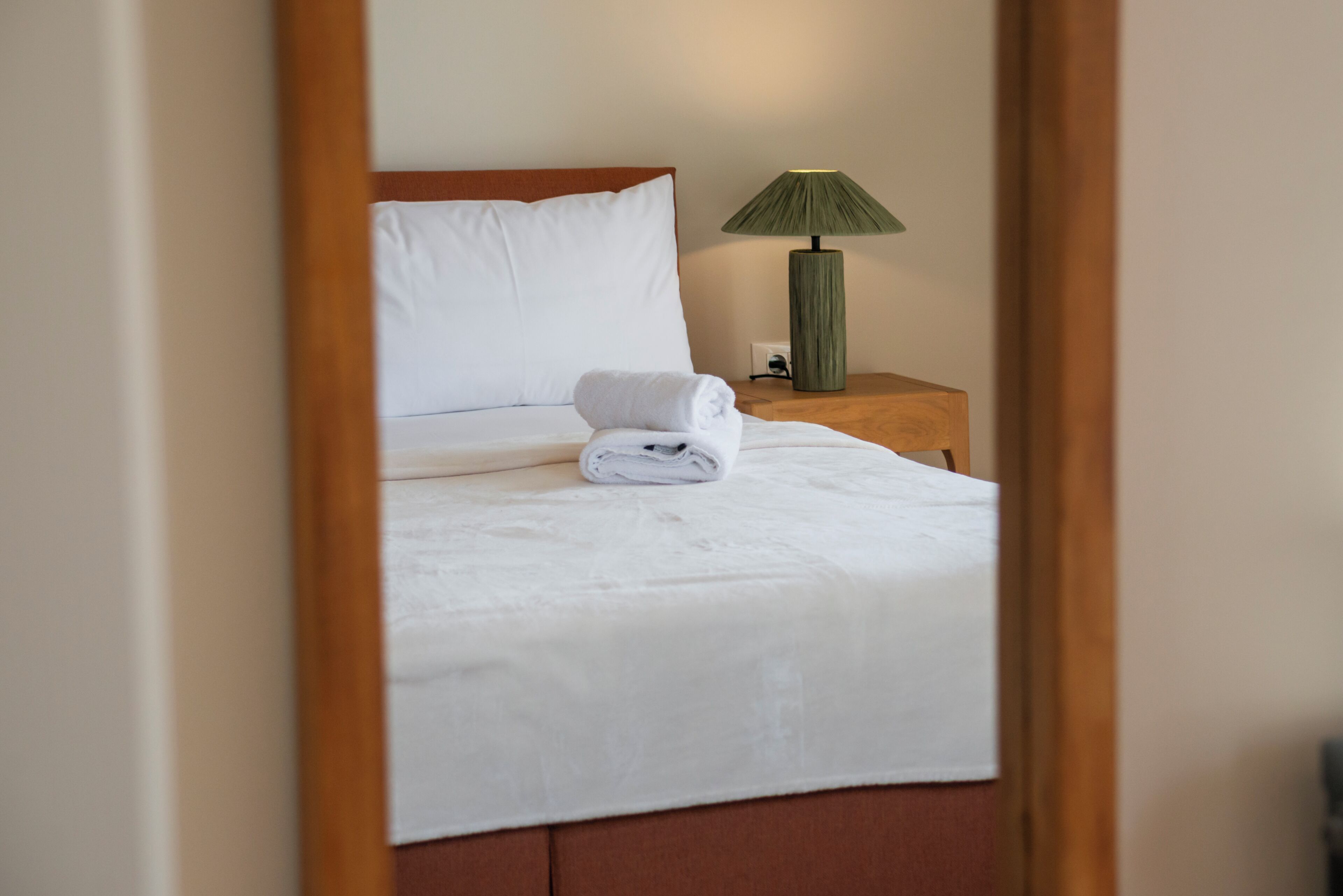 Un lit bien fait, avec du linge de lit blanc et une serviette pliée, une lampe verte posée sur une table de chevet en bois, et un miroir qui reflète la pièce.
