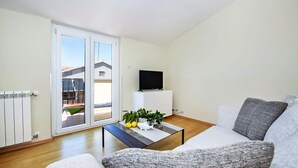 Una sala de estar moderna con un sofá, una mesa de centro con objetos decorativos, un televisor sobre un mueble bajo y una puerta corrediza de vidrio que da a un espacio exterior.