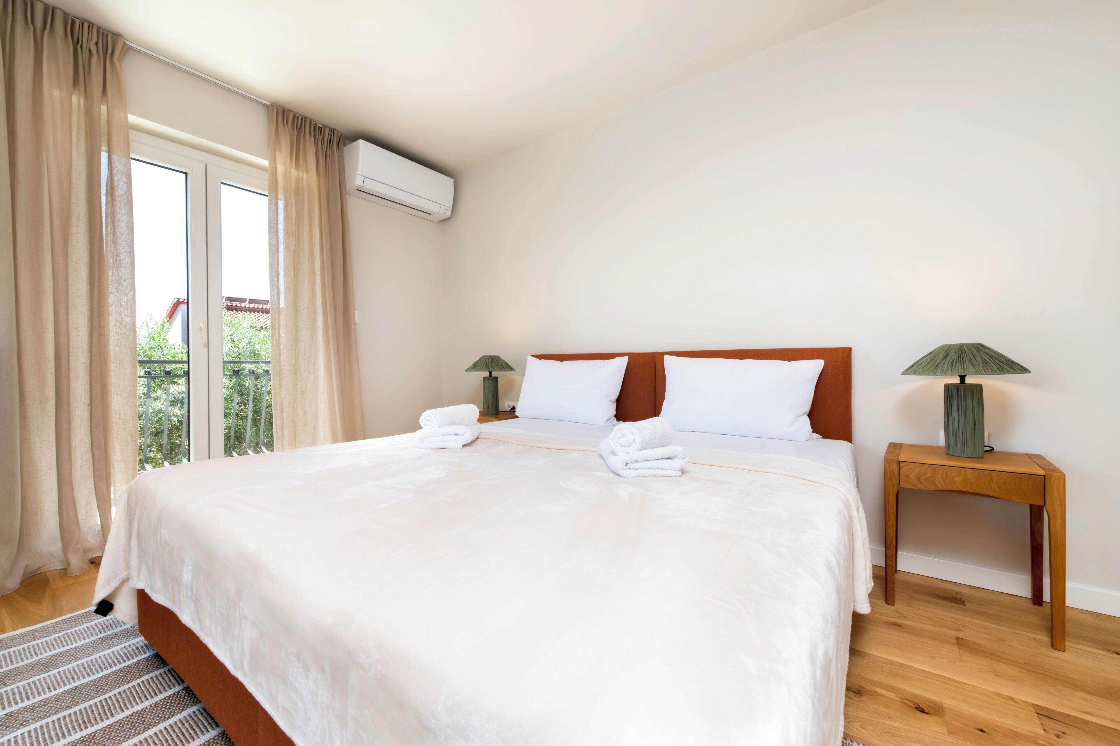 A hotel room with a large bed, two bedside tables, a window with curtains, and an air conditioning unit.