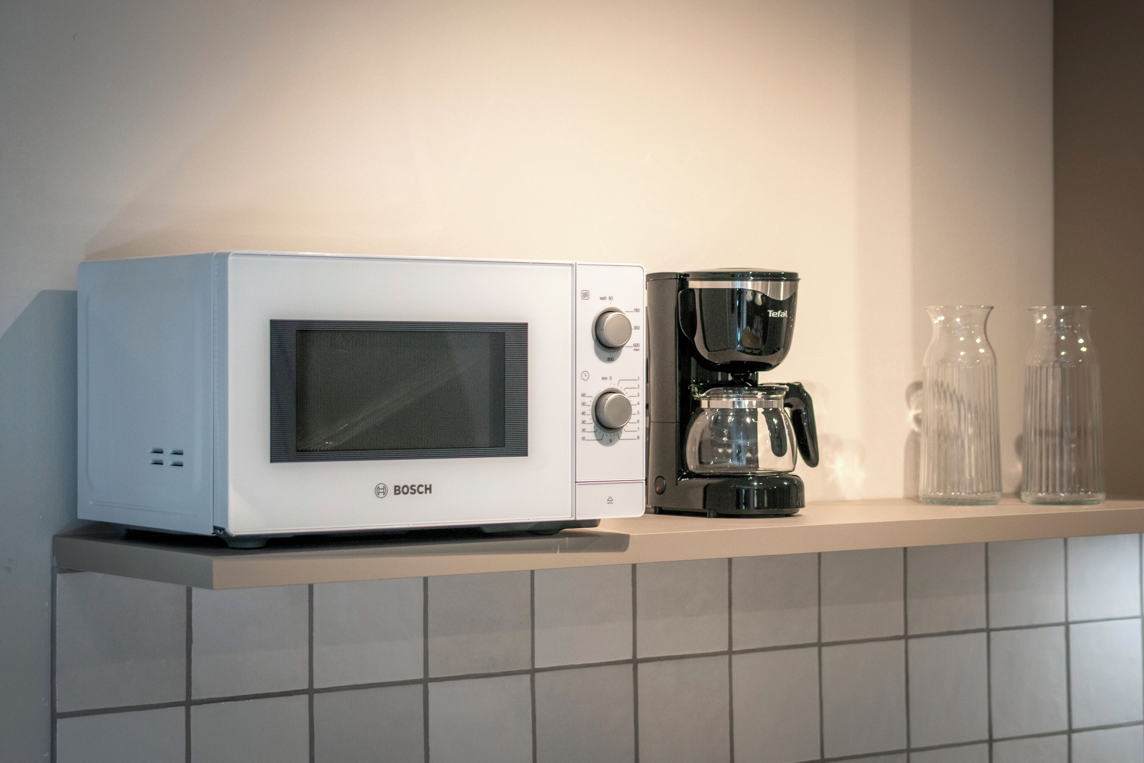 Eine Küche mit einer weißen Bosch-Mikrowelle, einer schwarzen Kaffeemaschine und zwei klaren Glasflaschen auf einem Regal.