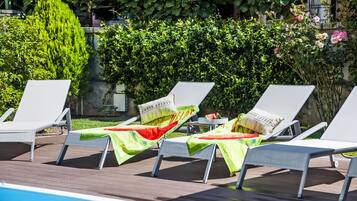 Zona de piscina con sillas reclinables blancas y una mesa, rodeada de abundante vegetación y un muro de piedra.