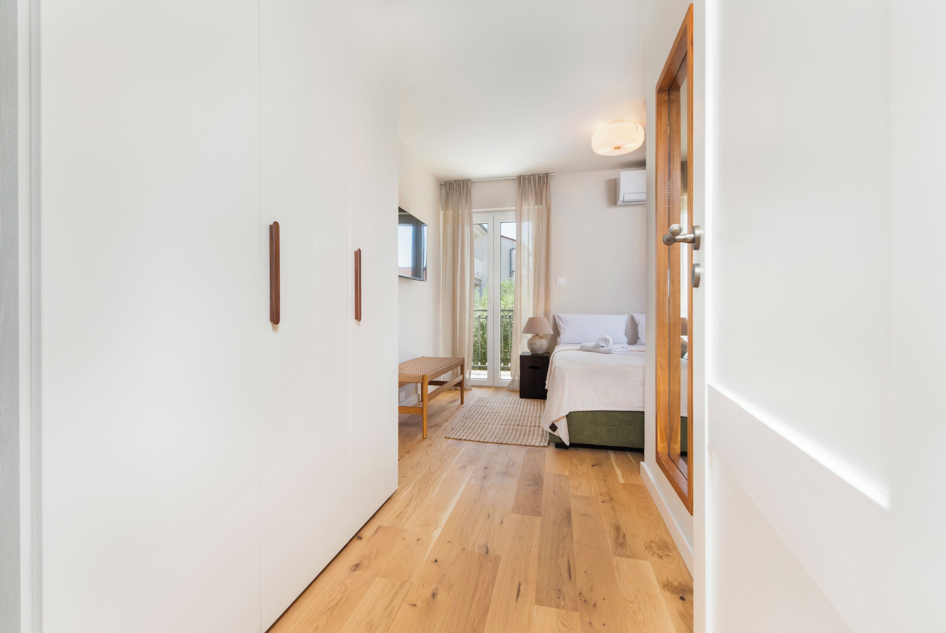 Un couloir mène à une chambre à coucher qui contient un lit, une table de chevet, une lampe et offre une vue sur l’extérieur.