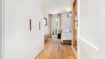 Un couloir mène à une chambre à coucher qui contient un lit, une table de chevet, une lampe et offre une vue sur l’extérieur.