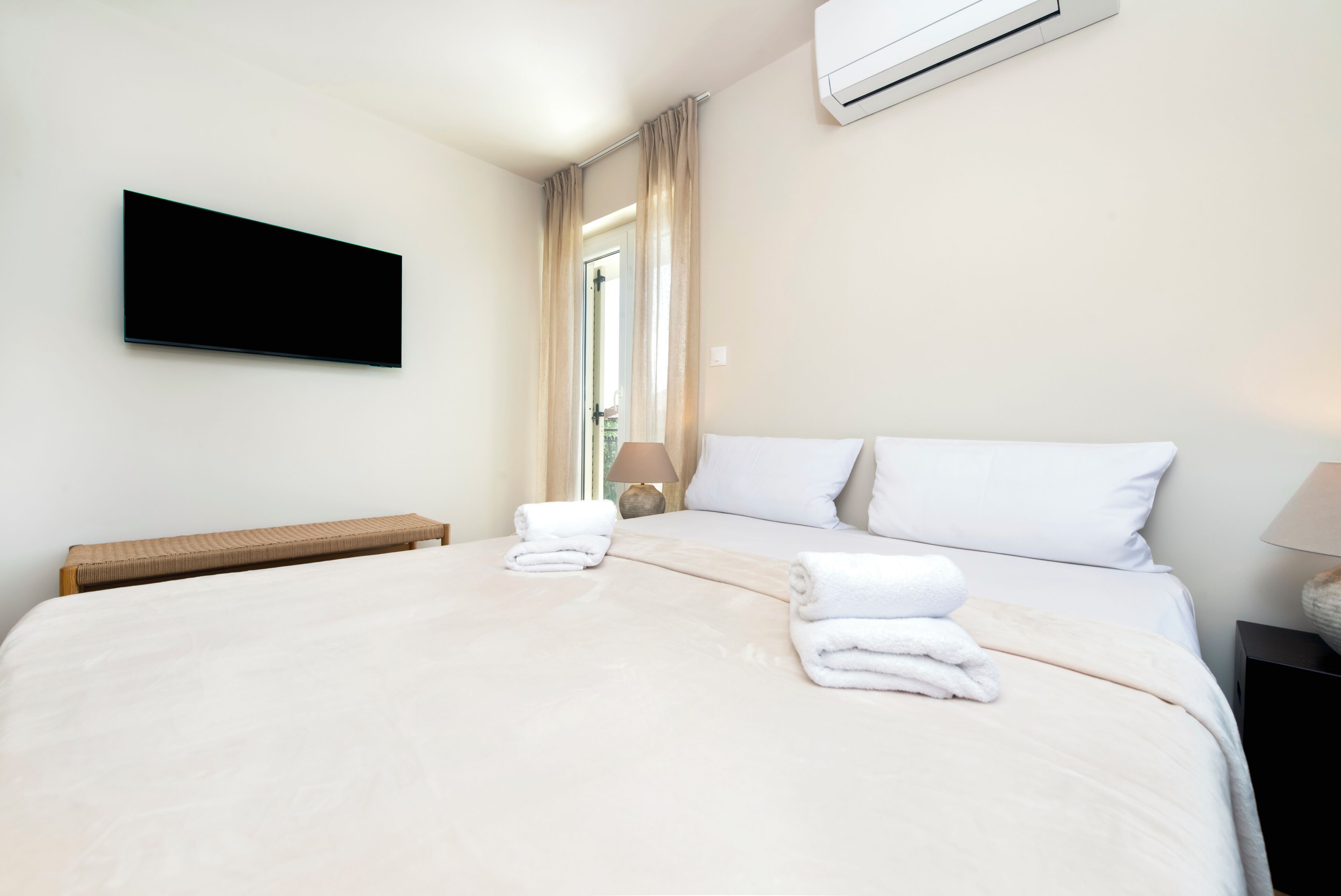 A hotel room with a bed, a flat-screen TV mounted on the wall, a bedside table with a lamp, and an air conditioning unit.