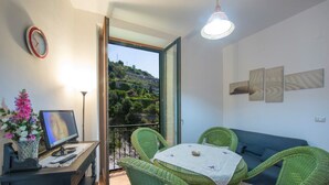 A balcony with a view of a hillside, a green wicker sofa, a small table with a tablecloth, a clock, and a framed picture on a desk. - Casa di mare, Exclusive Property on the beach, private terraces, stanning views (Maiori)