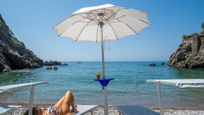 A beach with a white umbrella, rocky cliffs, clear blue water, and a person lying on a lounge chair. - Casa di mare, Exclusive Property on the beach, private terraces, stanning views (Maiori)