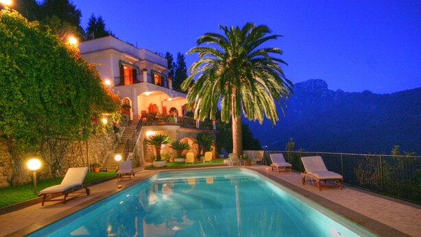 A poolside area with lounge chairs, a palm tree, and a building with multiple balconies. - Lo sguardo sul mare, Luxury Ravello Villa, Private Pool, Great View, parking (Ravello)