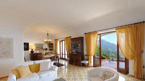 A spacious living room with a white sofa, a dining area with a wooden cabinet, and a view of mountains through large windows. - Lo sguardo sul mare, Luxury Ravello Villa, Private Pool, Great View, parking (Ravello)