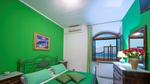 A room with a green wall, a bed with green bedding, a nightstand, a dresser, a mirror, a window with a view of the sea, and a painting on the wall.