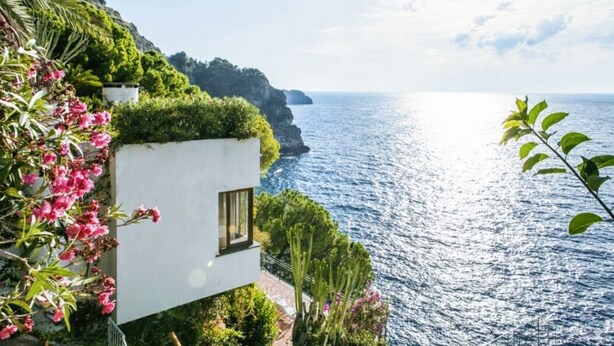 Um edifício costeiro com vista para o mar, rodeado por vegetação e flores.