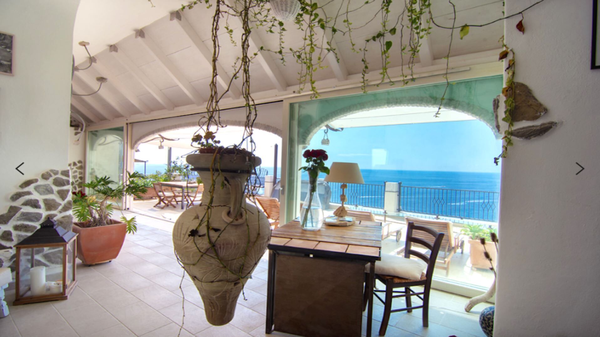 Une terrasse ensoleillée avec un mur en pierre, un vase suspendu et une vue sur la mer.