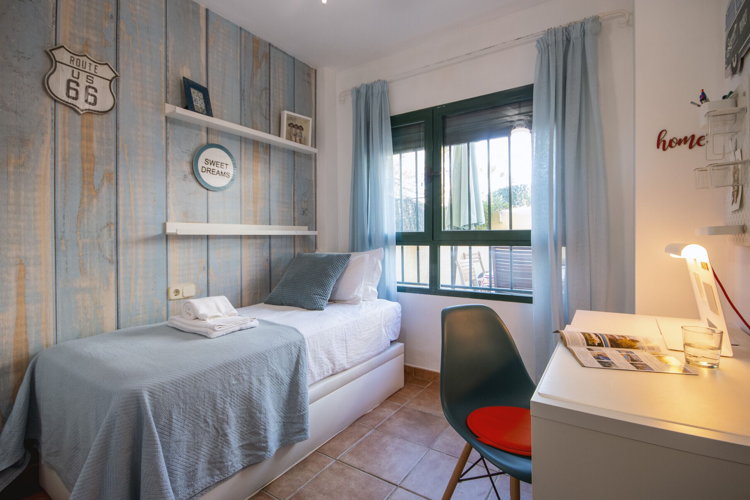 Une chambre à coucher avec un lit, un bureau avec une chaise, une fenêtre avec des rideaux et un mur orné d’un panneau et d’une étagère.