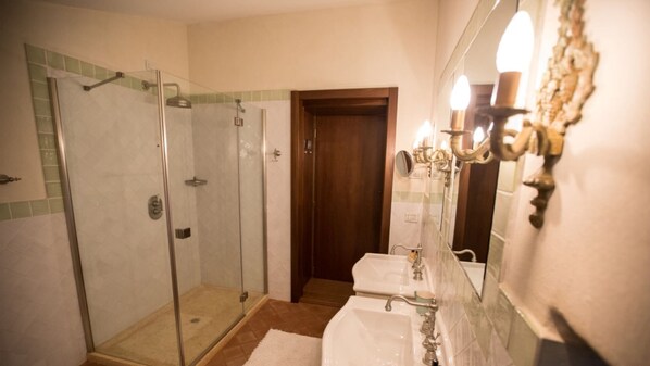 A bathroom with a glass-enclosed shower, two sinks, a mirror with wall sconces, and a wooden door.
