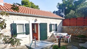 A stone building with a tiled roof, a door, and windows with shutters. There is a table and chairs outside.