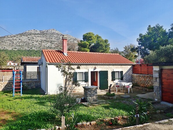 A small house with a red-tiled roof, a stone chimney, and a garden with a wooden fence and a ladder.