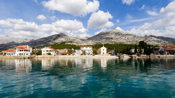 A coastal town with buildings along the waterfront, a clear sky, and mountains in the background.