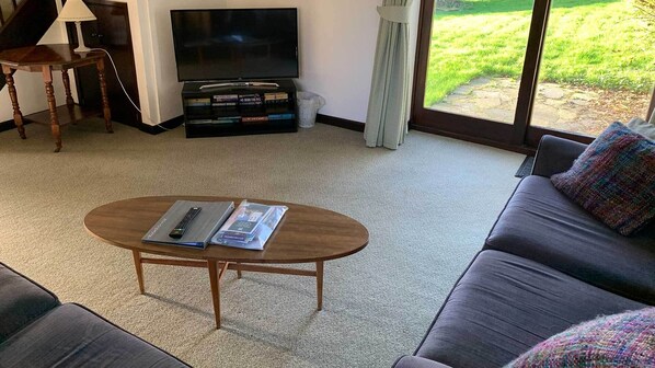 Un salon comprenant un canapé, une table basse avec une télécommande et des magazines, un meuble pour la télévision avec un téléviseur à écran plat, et une fenêtre donnant sur un espace verdoyant.