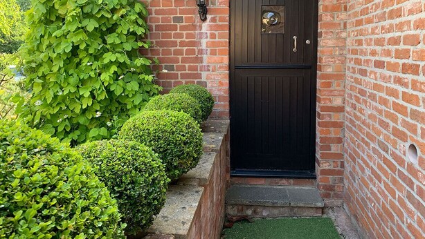 A black door with a brass knocker, a brick wall, and a small garden with neatly trimmed bushes. - Hayloft Cottage - Devon (Sidmouth)