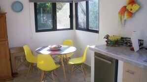 Un coin repas avec une table ronde et quatre chaises jaunes, une cuisine équipée d’un lave-vaisselle et une horloge murale.