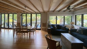 Un amplio salón con vigas de madera, una mesa de comedor con sillas, un sofá y grandes ventanales que dan vistas a árboles.