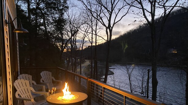 Una terraza con sillas Adirondack, una mesa redonda con una chimenea y vistas a un río y colinas.