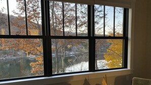 Una habitación con un ventanal grande que da a un lago y árboles otoñales. Hay una mesita con un barco modelo y una silla.