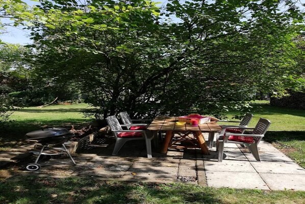 Ein Essbereich im Freien mit Holztisch und -stühlen, einem Grill und einem Baum, der Schatten spendet.