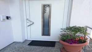 Uma porta branca com um painel de vidro vertical, uma maçaneta de metal e um tapete preto na entrada.