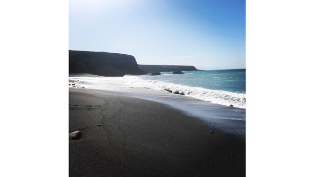 Ein Strand mit schwarzem Sand, Wellen die aufschlagen und eine Klippe im Hintergrund.