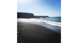 A beach with a black sand shore, waves crashing onto the beach, and a cliff in the background.