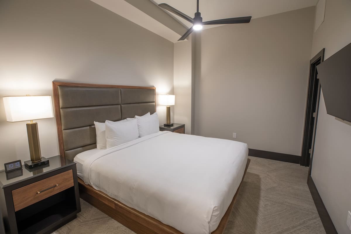 A hotel room with a large bed, two bedside tables, a nightstand, a ceiling fan, and a wall-mounted TV.