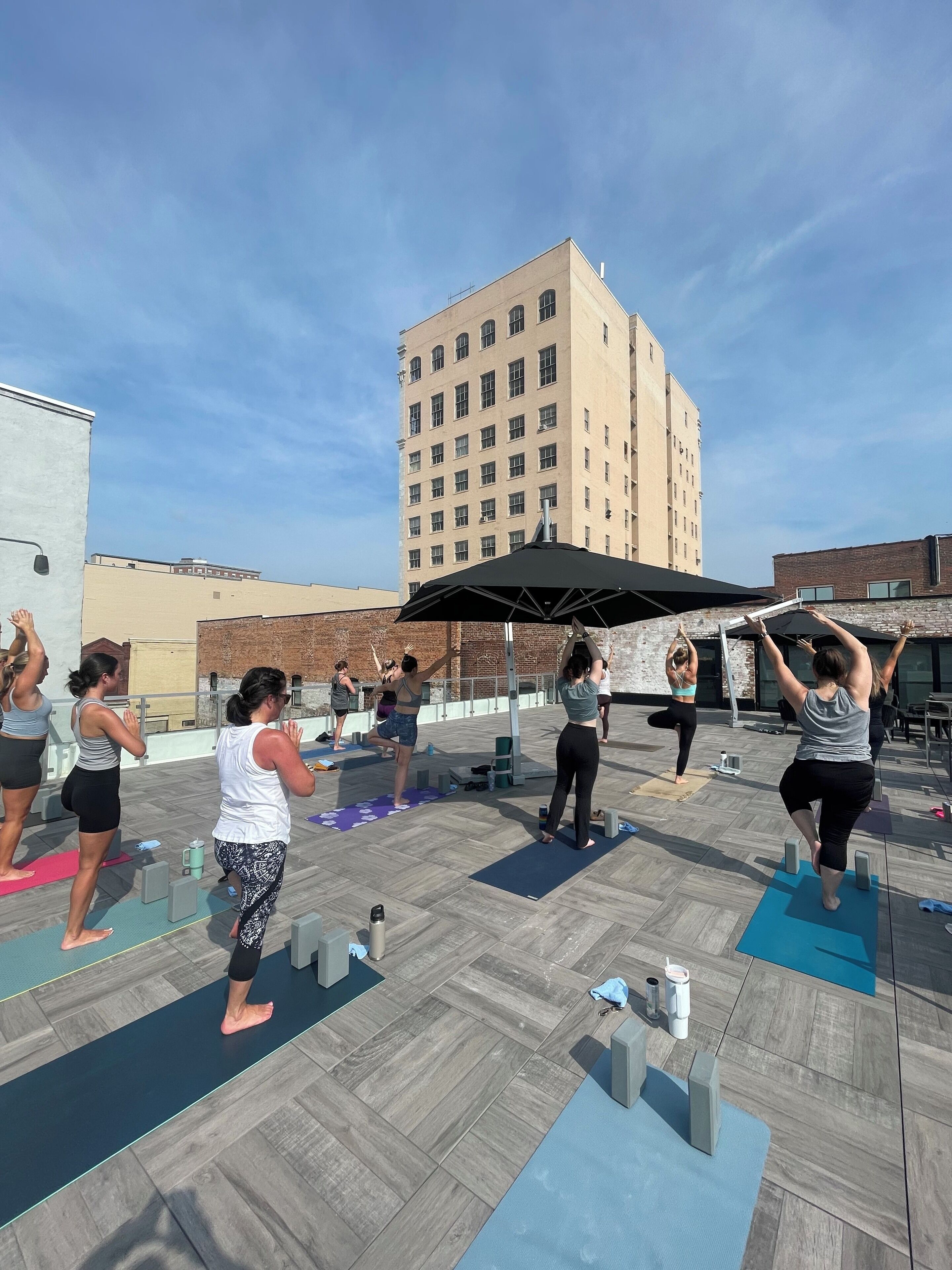 Um grupo de pessoas praticando yoga em tapetes ao ar livre em ambiente urbano, com edifícios ao fundo.