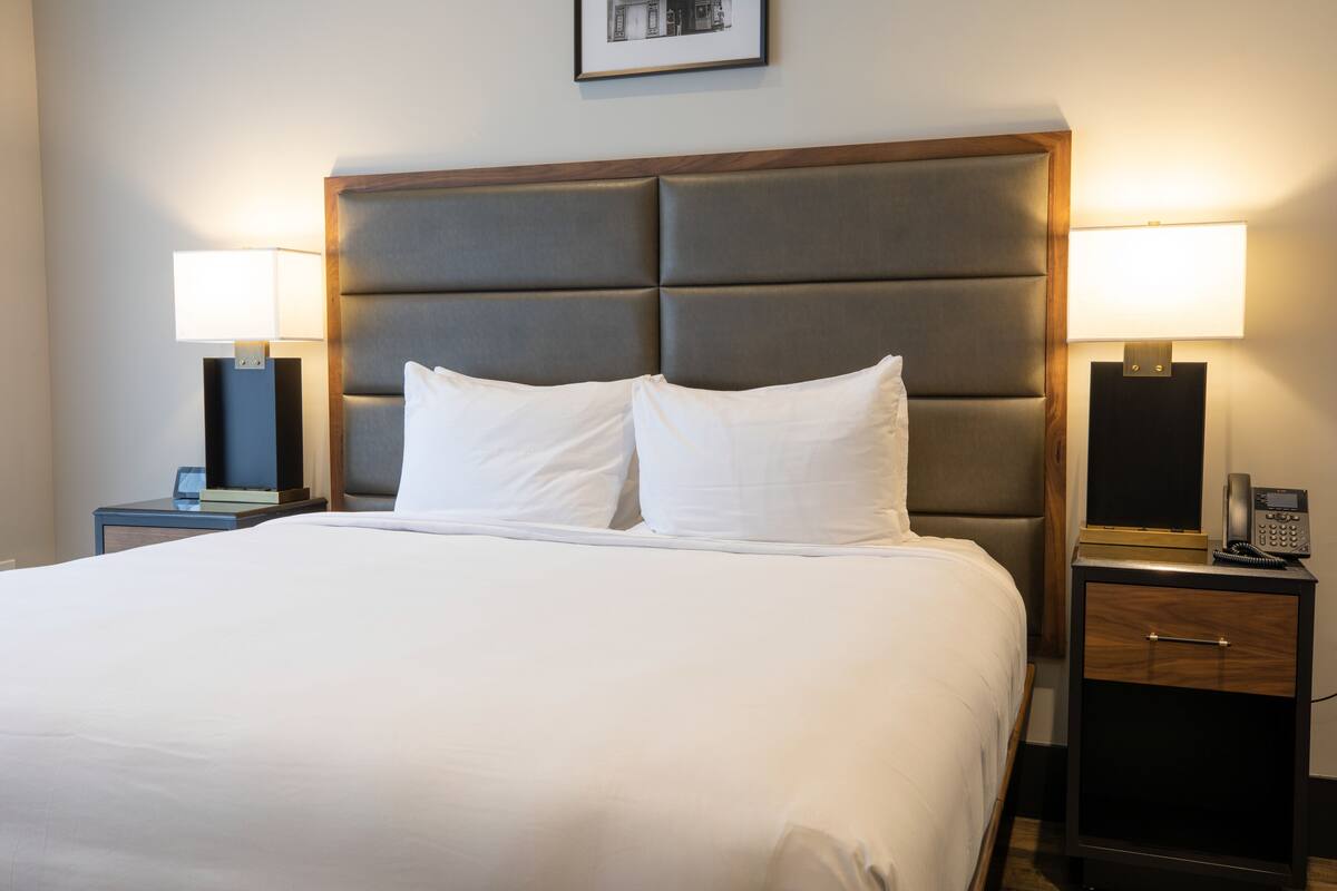 A neatly made bed with white linens, two bedside lamps, a nightstand with a phone, and a framed picture on the wall.