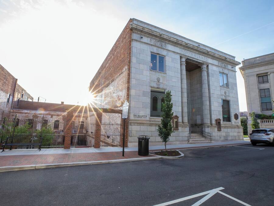 A historic building with classical architecture, a paved sidewalk, a street with a car, and a bench.
