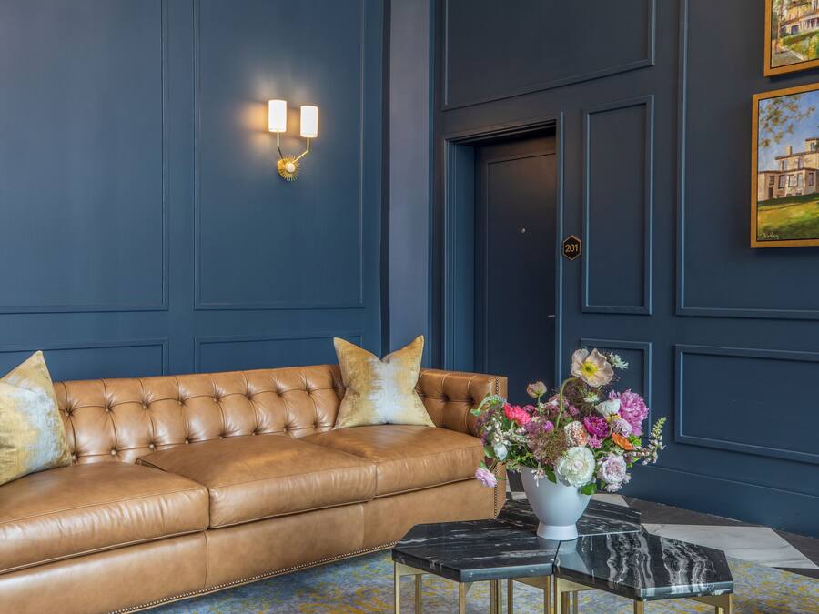 A room with a brown leather sofa, a marble coffee table, a wall-mounted light fixture, and a door with a number plate.