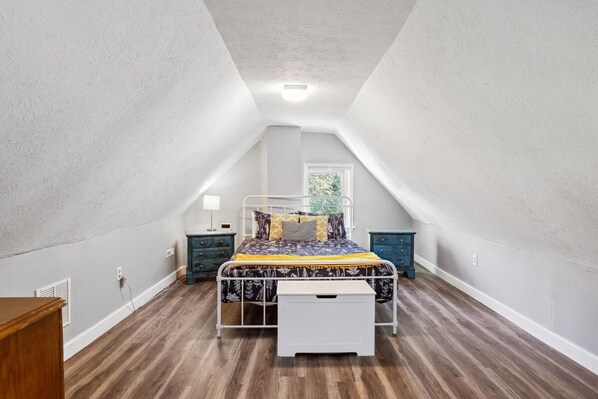 A bedroom with a sloped ceiling, a bed with a metal frame, a bedside table, a nightstand, and a window.
