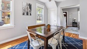 A dining area with a glass-top table, white chairs, a chandelier, and a blue patterned rug.